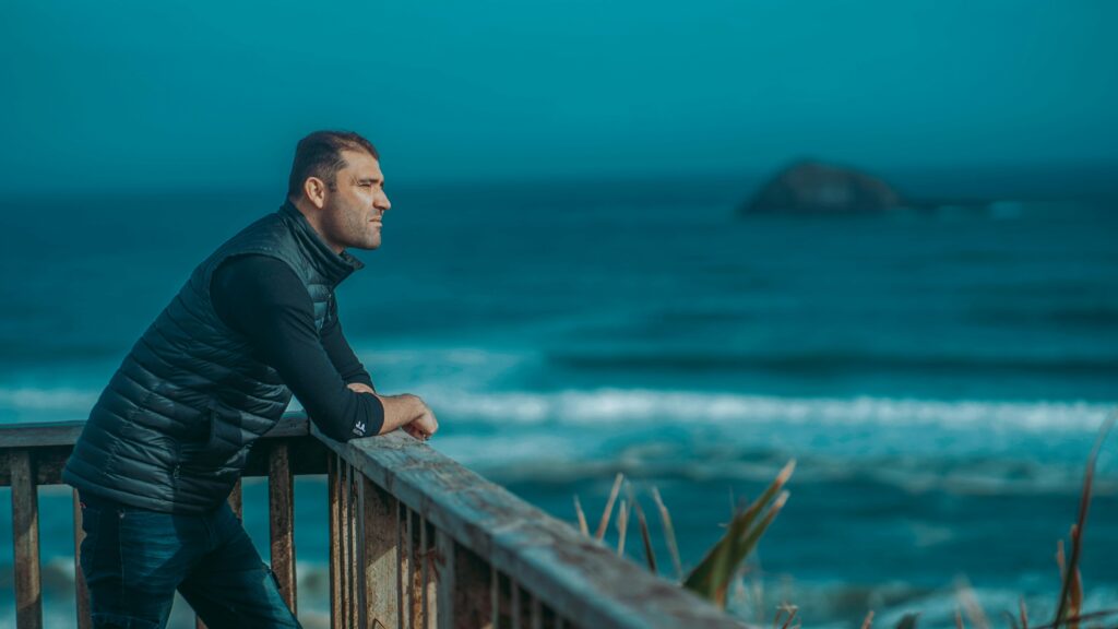 Picture of a man leaning against a worn wooden rail looking out over the ocean. It is late afternoon and you can see the waves breaking on the shore